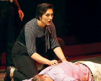 Antonia, in black blouse with black and white striped shoulders and sleeves to elbows and a bow on the collar and black pants kneels next to Caesar's bloody body; she's holding a knife in her righ hand and looking up at someone beyond the body