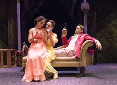 Hermia in a peach gown sits on the lap of Helena in a yellow gown, who sits on the edge of the chaise lounge as Lysander in salmon jacket leans back on the chaise lounge. The women have champagne glasses, Lysander the bottle. 