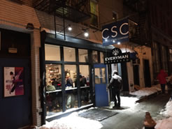 CSC marque over a glass-fronted section of building, a man opening the blue front door with lobby full of people inside the windows, snow on the edges of the sidewalk, and poster of Twelfth Night to the left of the windows. At night