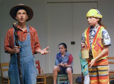 Corin in orange checked shirt, blue overalls, wide-brimmed hat and holding a curvey carved walking stick, Touchstone in yellow knit cap, blue floral shirt, bright orange and red vest, and horizontally multi-color striped pants, and holding a small brown whisky jug.