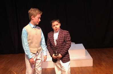 Bassanio in tan vest, light blue shirt, white slacks and a tie listens to Gratiano in a burgundy with checked jacket and white slacks, a flask in his hand.