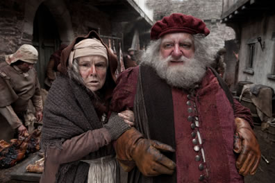 Mistress Quickly covered in raged clock holds onto Falstaff's Arm, who is in a medeivel lords jacket and cap with white beard and leather gloves in an narrow street with commercial activity in front of the houses