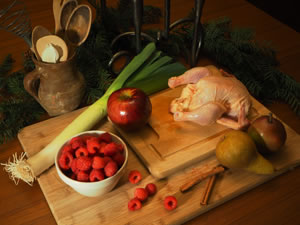 On two stacked cutting boards are a bowl of raspebrries, an apple, a leek, two cinamon sticks, two different pairs, and an uncooked game hen, with an old mug of wooden utensals and greenery in the background