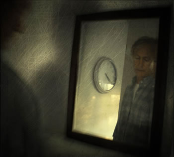 Reflection of a man in checkered work shirt in a mirror, he's looking down, and a clock on the wall is beside him in the reflection: time is 4:20; the shadow of his shoulder is to the left of the image, and the whole has a scratchy veneer