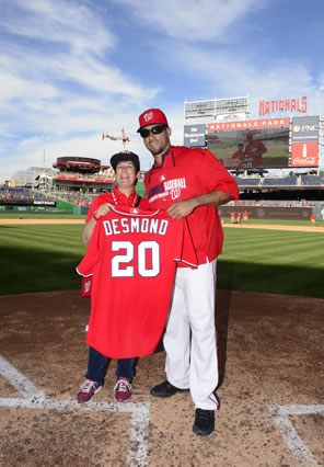 Desmond in red W basball cap, sunglasses, windbreaker and white uniform hat next to big-smiling Sarah in baseball cap, red shirt, blue jeans, and spider sneakers, both holding the red baseball jersey with "DESMOND 20" on the back. This is at home plate with the baseball field, scoreboard, and Red Porch cafe and beautiful blue skies with whispy clouds in the background.