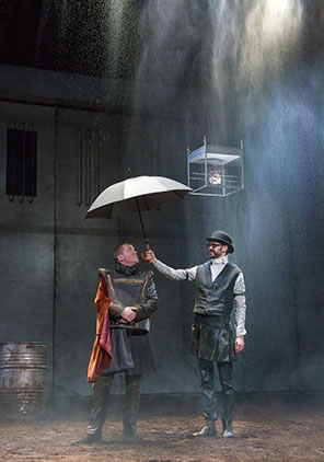 Production photo of Catesby and Hastings standing in the rain, Catesby, wearing a black bowler hat, black vest and white clerk's shirt, holding an umbrella over Hastings, who's in Elizabethan jerkin and skirt with a red cape on his shoulder.