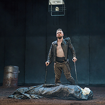 Production photo of Richard, in black unbuttoned Renaissance jacket and bround pants, leaning on his two crutches, standing by the shrouded body of Henry VI. The floor is dirt, a barrel is in the background, and a skull is suspended in a glass box hanging from the ceiling.