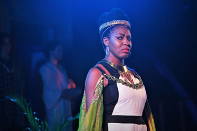 Prince Escalus in a classic white gown with black trimming, a headband crown, a green cape with jeweled chain around her shoulders and long earrings shows a frown of grief in her expression
