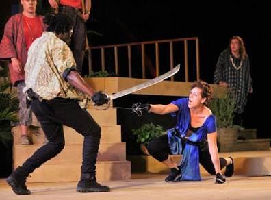Mercutio in gold and white shirt, black pants and boots with a sabre in his right hand and sword in his left parries with Tybalt, in blue toga, black braw and yoga pants kneeling on the ground with a sword thrusting in her right hand and dagger in her left. In the background other characters watch on a sets of wood steps and balconies.