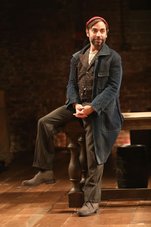 Feste in dirty blue overcoat, brown striped pants, brown dotted vest, and red knit cap sits on the edge of a large wooden table.