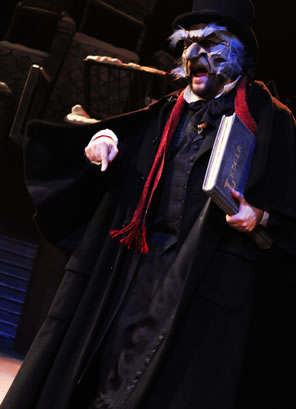 Scrooge in Victorian overcoat and eagle-like mask, carrying a ledger and angrily pointing his finger down.