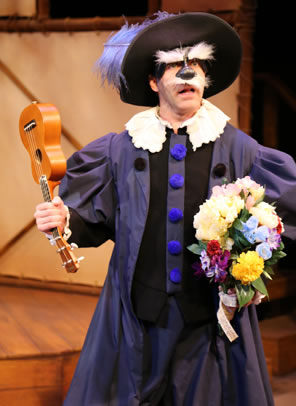 Il Dottore wearing a purple rennaissance robe with white frilly coller and huge blue buttons, a large-brim hat with purple feather, a mask featuring a black nose and huge white fluffy eyebrows and mustache. He's holding a ukelele by the neck in one hand and a bouquet of flowers in the other.
