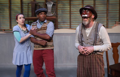 Harpagon in checkered pants, suspenders, gray checked vest, and dingy long-sleeved undershirt plus fodora hat and brown mask with a white mustache stands to the right while Elise in blue schoolgirl dress with white tights and Cleante in red pants, brown striped-patterned seater vest and blue shirt plus dog cam clasp each other in shock over what their father is saying. Chairs are on a platform in the background in front of a slatted wood wall. 