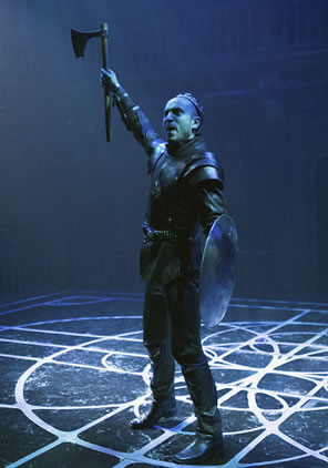 Richard in short skirt with shield in one hand and battle axe raised in the other on the stage in a rainy-quality light