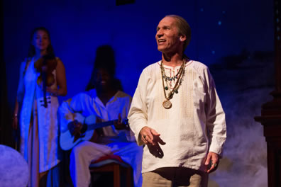 Gower wearing a natural linen tunic and several beaded necklaces and a medallion, with a violinist and guitarist in white in the background