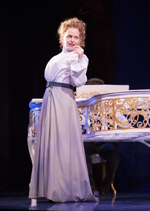 Tonya Beckman in simple Victorian-era white blouse and gray floor-length skirt stands next to a gold-etched white piano