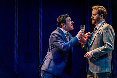 Leontes in blue suit and holding a champagne glass leans into and jabs his finger at the chest of the listening Camillo in gray three-piece suit.