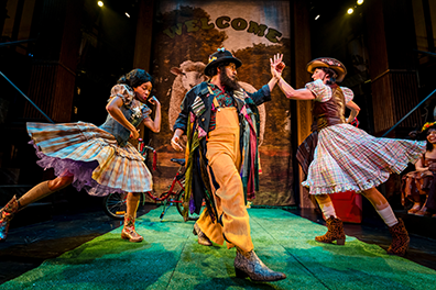 Autolycus in rustic hat, beard, multicolored coat over orange coverals dances with two women in peasant dresses on a green-grass stage with a large "Welcom" poster featuring a sheep on the back walla. 