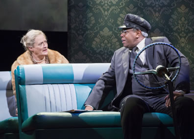 Daisy with gray hair back in a bun and wearing a mink stole with pearls around her neck in the back seat of the car, featuring shades of blue vinyl upholstery, with Hoke in three piece dark gray chauffeur uniform and cap turned around as they talk to each other.