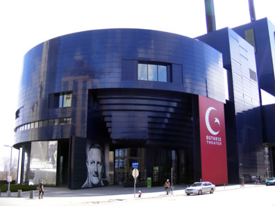 Deep blue building with curcular facade and two tall blocks further down, each with what appears to be smokestacks, a photo of Tyrone Guthrie next to the door, and a large red signe with a moon and star and "Guthrie Theater" covering one wall.