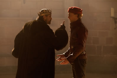 Henry in long fur robe and crown sticks a finger up at the face of Hal in red short jacket and red cap, his hand out in ront of him. Stone wall in the background.