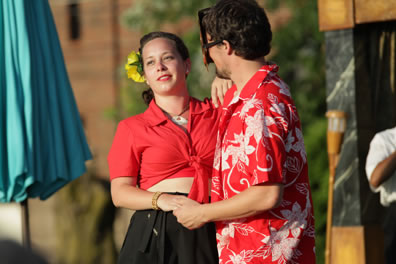 Beatrice in red shirt tied up to reveal a bare tummy above a black dress, a yellow flower in her hair, holds hands and has her other hand on the shoulder of Benedick as the dance, he in a red and white floral pattern shirt and wearing a mask. A big, blue beach umbrella is to the left.
