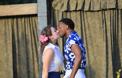 Hero, with white blouse tied up to reveal her bare midriff oer a blue dress and pink flower in her hair, kisses Claudio, wearing a blue and white flower print shirt and white pants; a surprised Claudio has his eyes wide open.