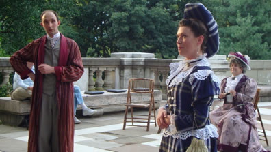 Algernon in three-piece gray suit and red striped dressing gown with hand on his and other hand across his stomach, Gwendolen in blue and white striped Victorian hat with lace trim and puffy hat, hands folded at her belly, Lady Bracknell in dress and straw hat with red bow sitting on a chair in the backround, where you see parquade floor, stone railing, and trees.