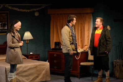 In the motel room, cast in winter jackets, Terrence (in orange and yellow uniform shirt) talks with Josh to one side while Ayelet watches to the side; you can see silver tinsel strewn over the bed.