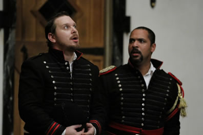 Iago and Othello on their knees, both in uniform, Othello's with shoulder bourds and gold and red braids, with his collar undone as he looks at Iago with concern