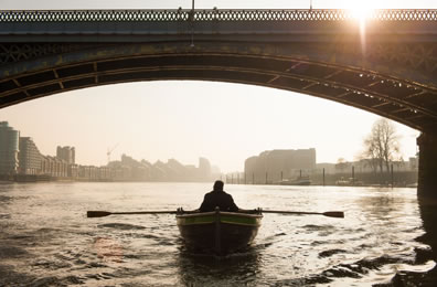 A figure in a row boat passes under a bridge on the Thames in London at sunset or sunrise (sun shining through the bridge railing)