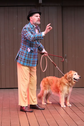 Launce wears a blue and black tartan jacket over tan pants, with bowtie and brown shoes and black bowler hat speaks with one hand gesturing in an OK sign, the other hand holding a thin red cane and a rope leash to the golden retriever standing stoicly looking off in the distance.