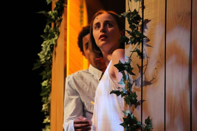 Juliet in a plain white nightgown and behind her Romeo in an Oxford shirt stand in a window with ivy vines on wood-paneled walls.