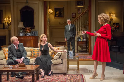 Mallonnee in three-piece suit sits next to Anna in long black dress and strapped high heels on couch, Chandler in background wearing gray business suit with left hand in pocket, Hester standing right, talking down to Mallonee and Anna, glass in left hand, making a point with right, wearing a knee-length red dress, white stockings and heels