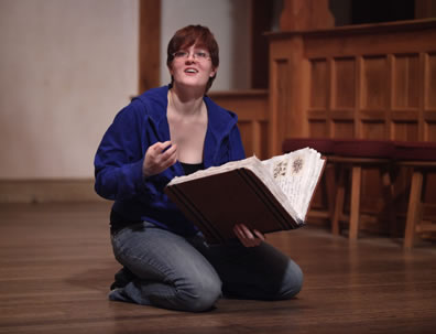Faustus kneels on the floor with a large book of spells in her hand as she talks out toward the audience
