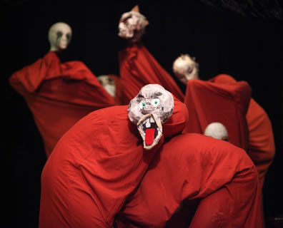 Characters in single red sheets and grotesque masks, the one in front with his mouth wide open and a large red tongue sticking out
