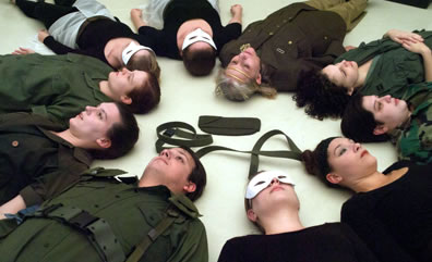 The cast lies on their backs on the ground in a circle, heads facing inward, the witches in white masks, olive green belt and a sky cap in the middle of the circle