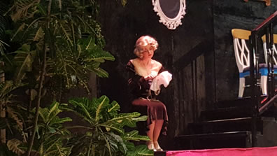 Ophelia in brown dress just below her knees works on needlepoint on a stage with red carpet, wood stairs, and green plants.