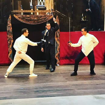 Hamlet in white with black glove holding foil faces off with Laertes in white fencing shirt and black pants, with Horatio in a suit and tie in the middle as referee