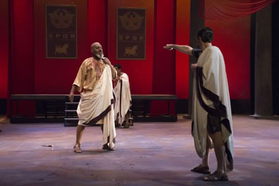 Caesar in bloodied toga and robe stands next to his dropped knife as Brutus, also in a toga and robe, stands with knife pointing toward Caesar. In the e background are the red-draped walls and SPQR posters