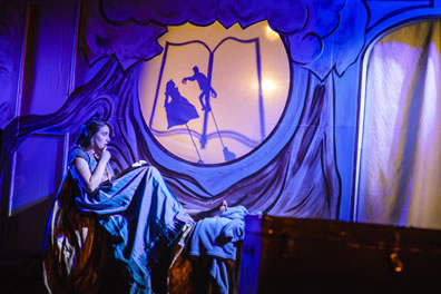 Imogen sitting in bed, wearing blue nightdress, book in her lap, and in window above her a shadow puppet of a man with long finges attackine a woman on an outline of a book. Iachimo's trunk is in the foreground of the photo