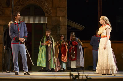 Benedick in pale blue army officers uniform with hands on hilt stands to one side across from Beatrice in yellow layered dress, while the other characters stand in the background in cloaks. 