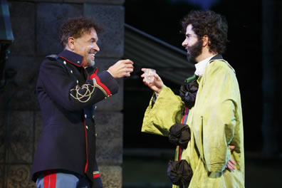 Don Pedro in dark blue army officers coat with red braids and light blue uniform pants with red piping holds a glass up to Benedick wearing a lime green clown costum with big puffy black buttons