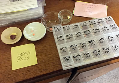 A pill organizer on a dresser top, with small dishes adjacent to it each containing pills, and a "Sarah's Pills" yellow Post-it note,