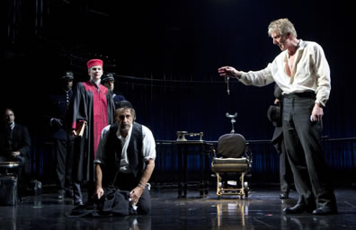 Production shot of trial scece: Antonio lroding over a kneeling Shylock with Portia looking on