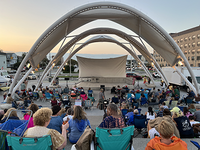 A view of the three-awning amphitheater looking from the boardwalk in toward the empty stage. People sitting in lawn chairs and on blankets, with buildings in the background and the sunset in the distance.