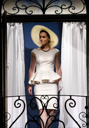 Siliva in tight white knee-length dress, a blouse that hugs the shoulder and bosom and spreads out in pleats at the waste, and a white wide-brimmed hat walks through the curtains of a wrought-iron balcony.