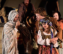 Production photo of Polixenes, scarf up to his face, talking with Florizel with his arms around Perdita as shepherds watch in the background.