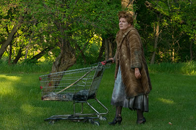 Production photo of Timon wearing a fur coat and pushing a shopping cart in a meadow with trees in the background