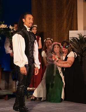 Claudio in dark green waistcoat, puffy white shirt, and pants and boots, stands center stage as a veiled Margaret kneels in the background with the other women crowded around her.